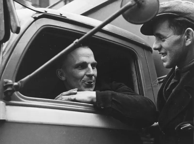 A truck driver leans on the open cab window and talks with a helper standing beside the truck.