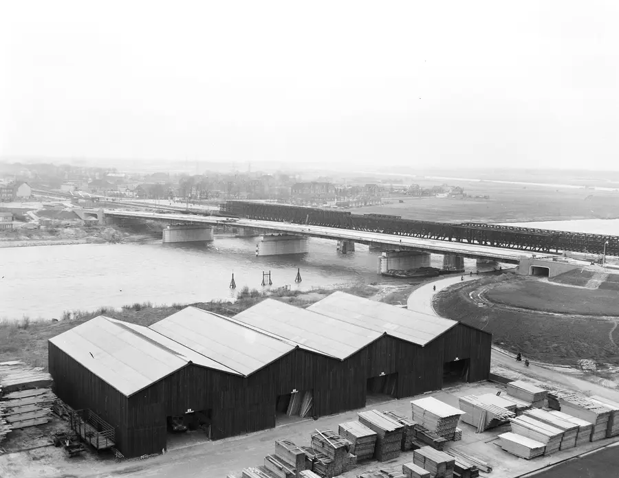 Seen from above: the new road bridge over the Maas at Venlo under construction, next to the old railway bridge, with timber sheds on the bank.